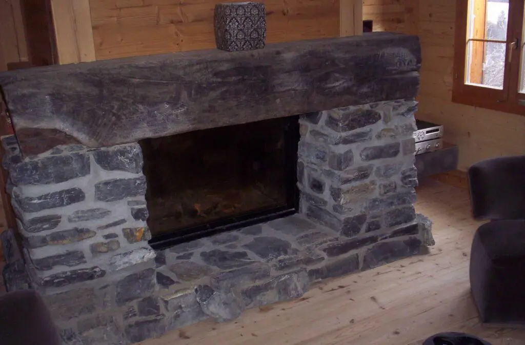 Rustikaler Steinkamin in einer Holzhütte, ergänzt durch einen Holzbalkenmantel und gemütliche Einrichtung.