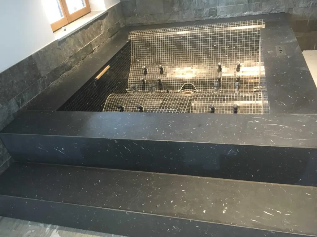 Moderner, unfertiger Jacuzzi mit dunklem Fliesenrand und Stufen in einem Innenraum unter einem Fenster.