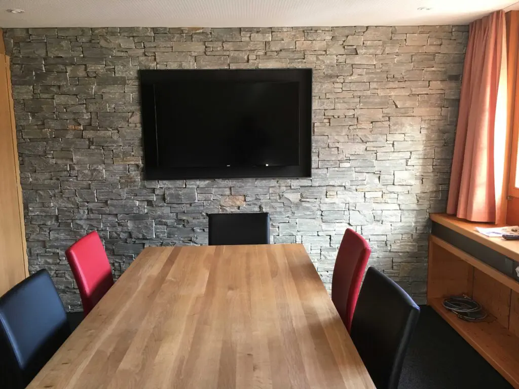 Moderner Konferenzraum mit Holztisch, Steinwand und Flachbildfernseher. Gemütliche Atmosphäre mit farbigen Stühlen.