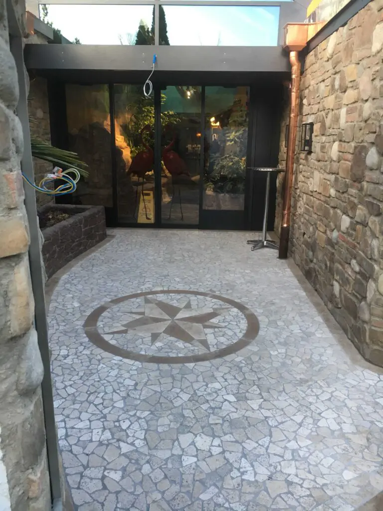 Steinterrasse mit Mosaikstern und Glasschiebetür, flankiert von Steinwänden, führt zu einem Innenraum mit Pflanzen.