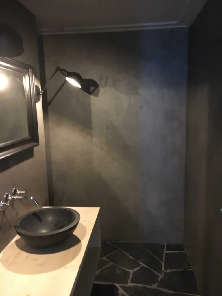 Modernes Badezimmer mit schwarzem Waschbecken, Betonwänden, heller Holzablage und stilvoller Beleuchtung.