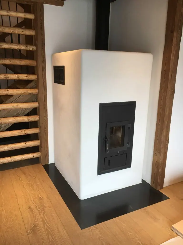 Moderner Kachelofen im Wohnzimmer mit Holzboden und Holztreppe im Hintergrund.