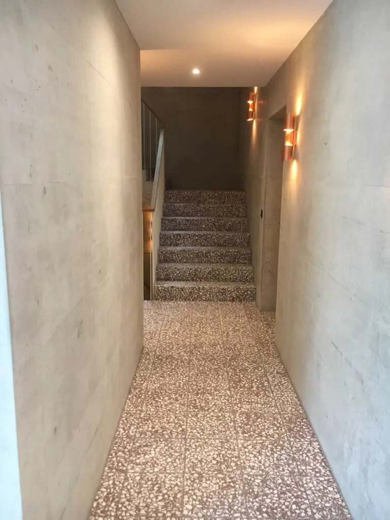 Moderner Flur mit Betonwänden und Terrazzo-Boden, Treppe und Wandleuchten im Hintergrund.