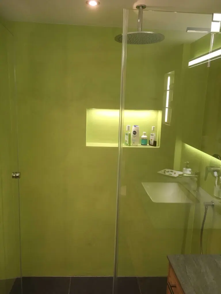 Grünes Badezimmer mit Glasduschwand, Regendusche, eingelassenem Regal und Waschbecken; modernes Design.