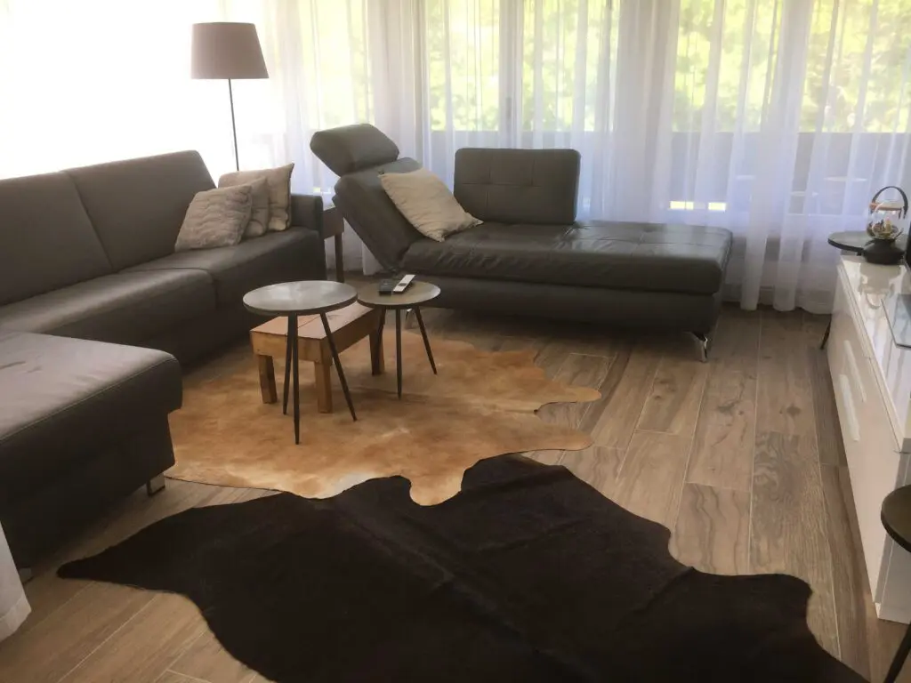 Modernes Wohnzimmer mit Ledersofas, Couchtischen und Teppich auf Holzfußboden, umgeben von großen Fenstern mit Vorhängen.