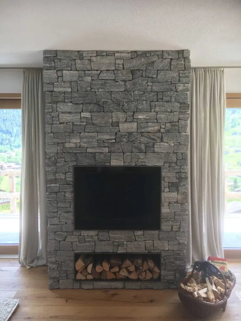 Moderner Kamin mit grauer Steinverkleidung und Holzlager, umgeben von großen Fenstern und Vorhängen im Wohnzimmer.
