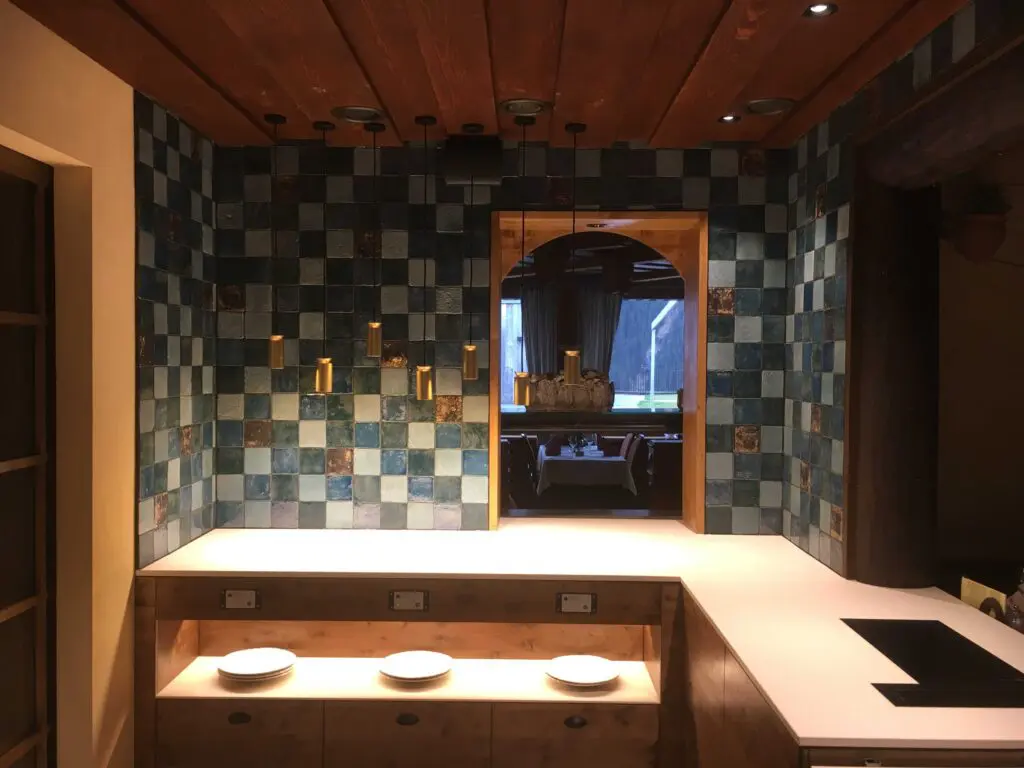 Moderne Küche mit blau-grauem Fliesenspiegel, Holzmöbeln und Hängelampen, Blick ins Esszimmer.