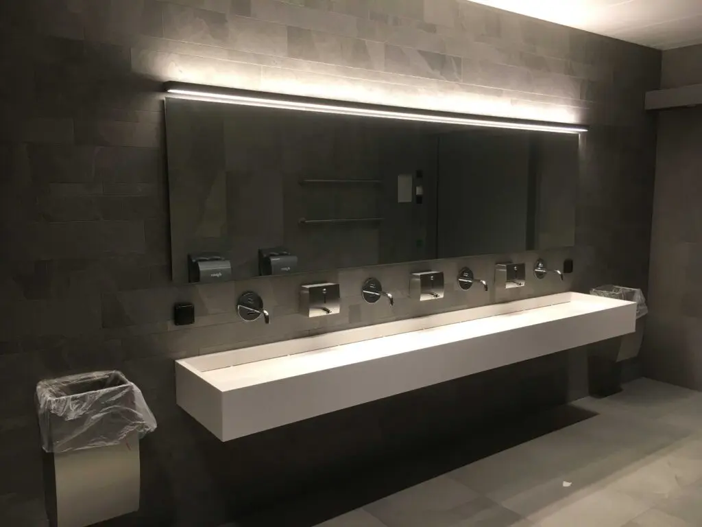 Moderne, stilvolle Toilette mit großem Spiegel, mehreren Waschbecken und Seifenspendern an grauer Steinwand.