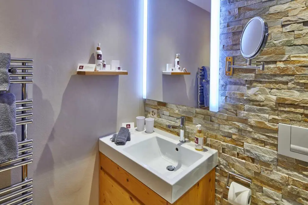 Modernes Badezimmer mit Waschbecken, beleuchtetem Spiegel und Steinwand, ausgestattet mit Handtüchern und Pflegeprodukten.